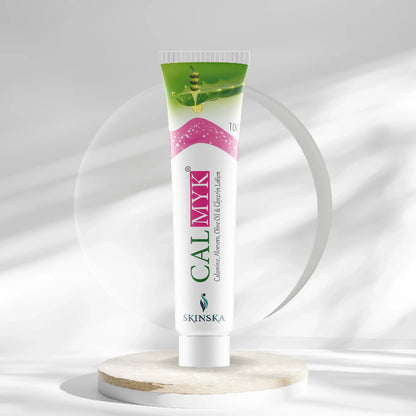 A tube of CALMYK ANTI-ACNE CREAM positioned upright on a wooden surface with a white background. The packaging is primarily white with green and pink accents, and the text indicates the product is for acne-prone skin.